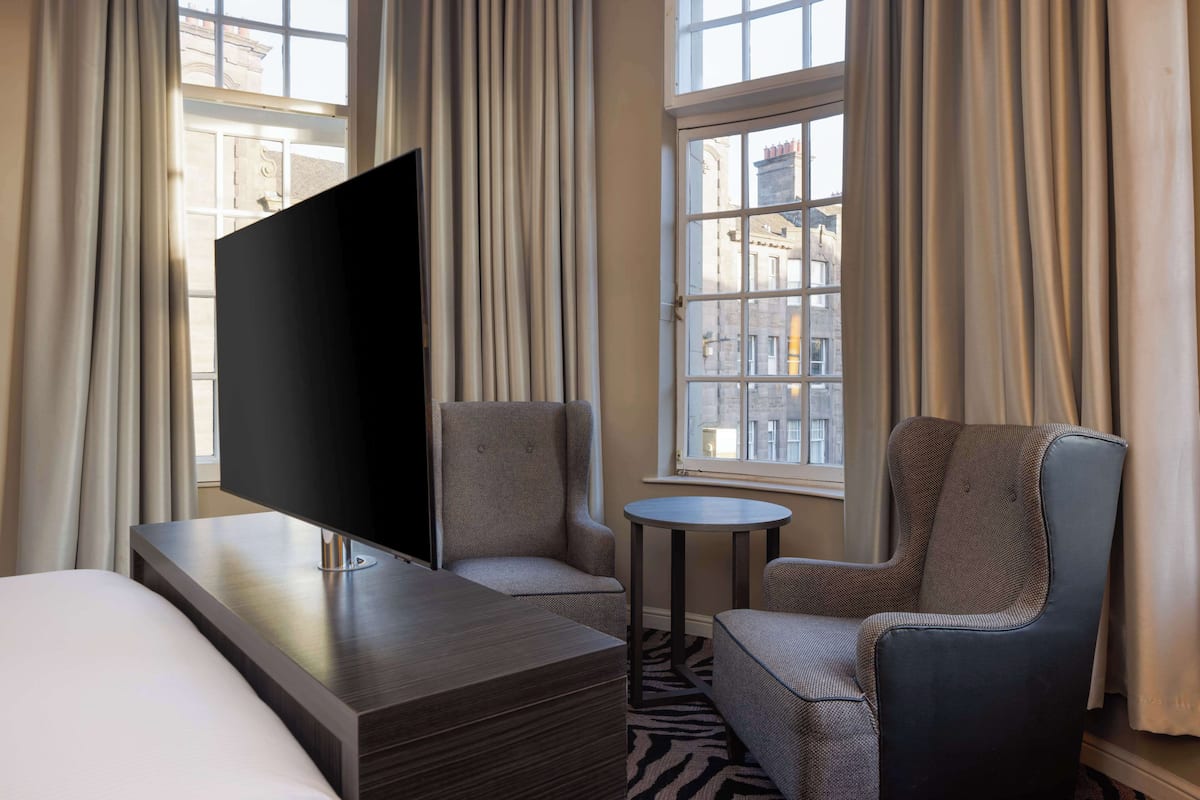 A hotel room with a bed, a TV, two armchairs, a small round table, and a window with curtains.