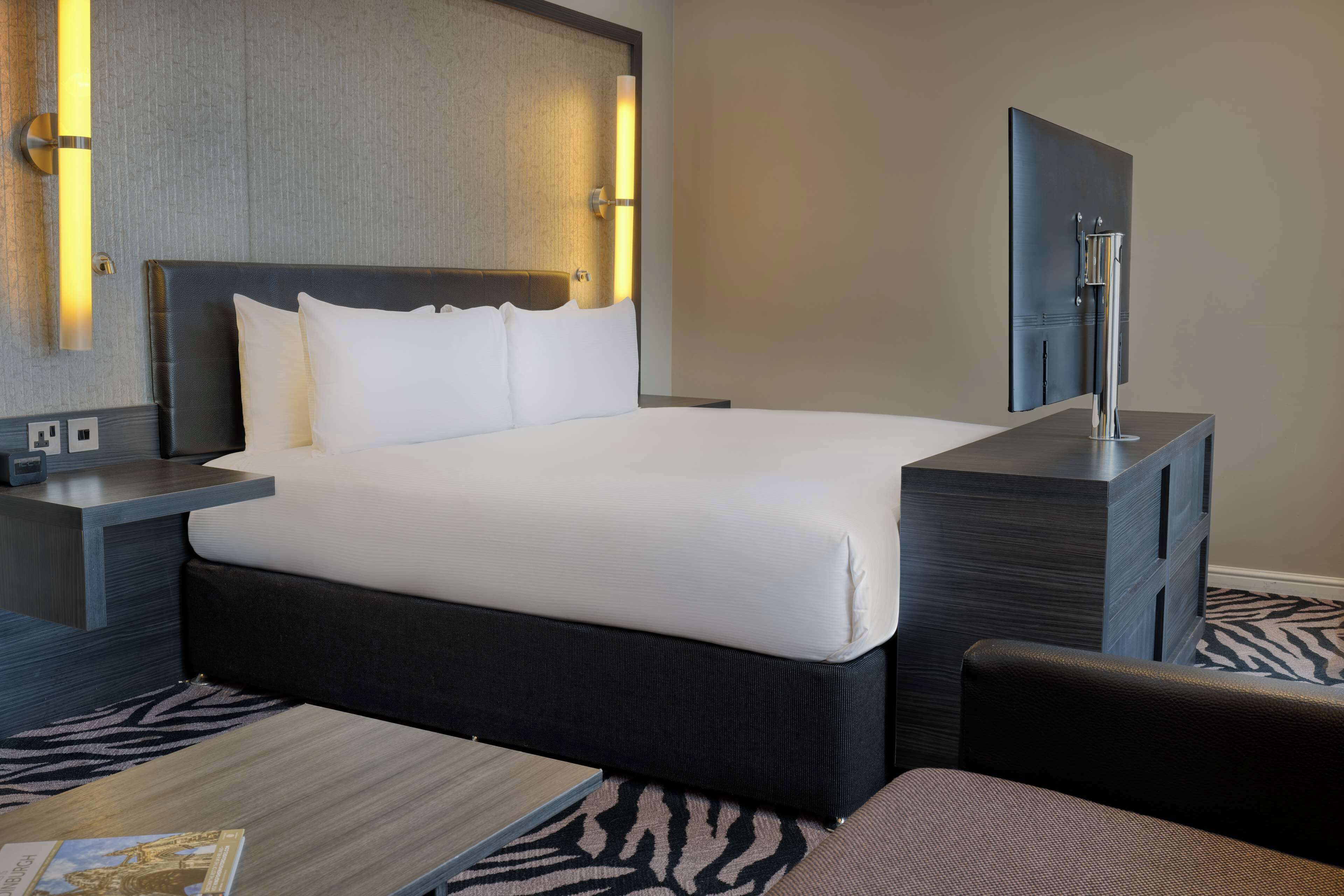 A modern hotel room with a large bed, a bedside table, a nightstand, a flat-screen TV, and a small seating area.