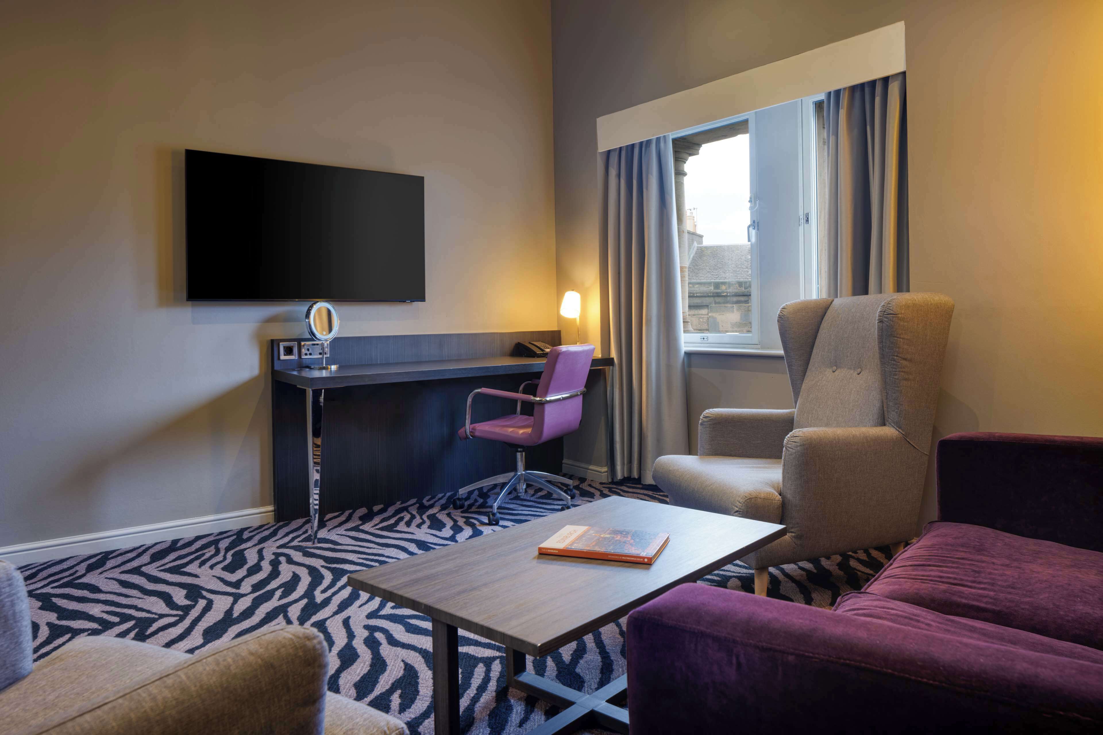 A hotel room with a flat-screen TV mounted on the wall, a desk with a chair, a sofa, and a large window with curtains.