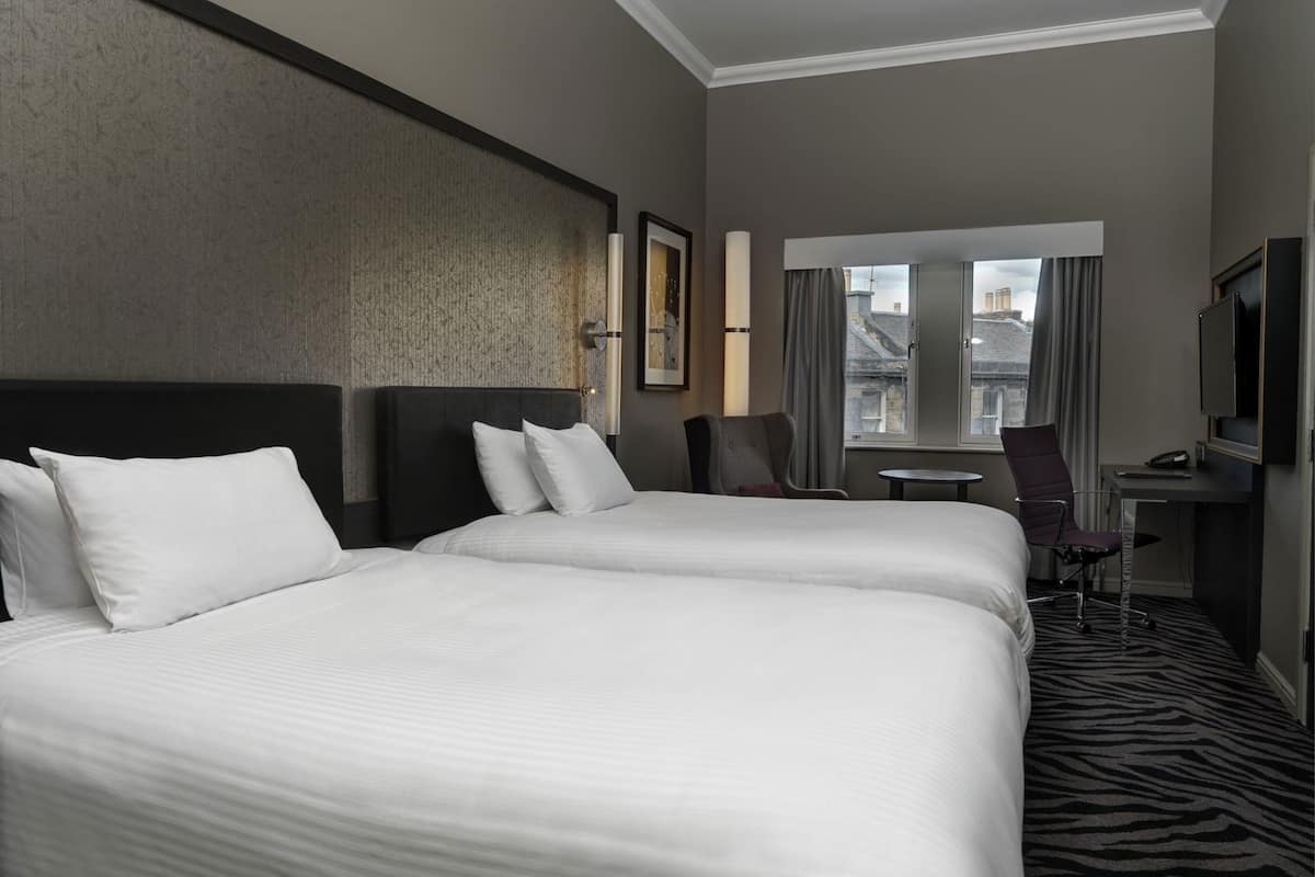 A hotel room with two beds, a desk, a chair, a TV, and a window with curtains.