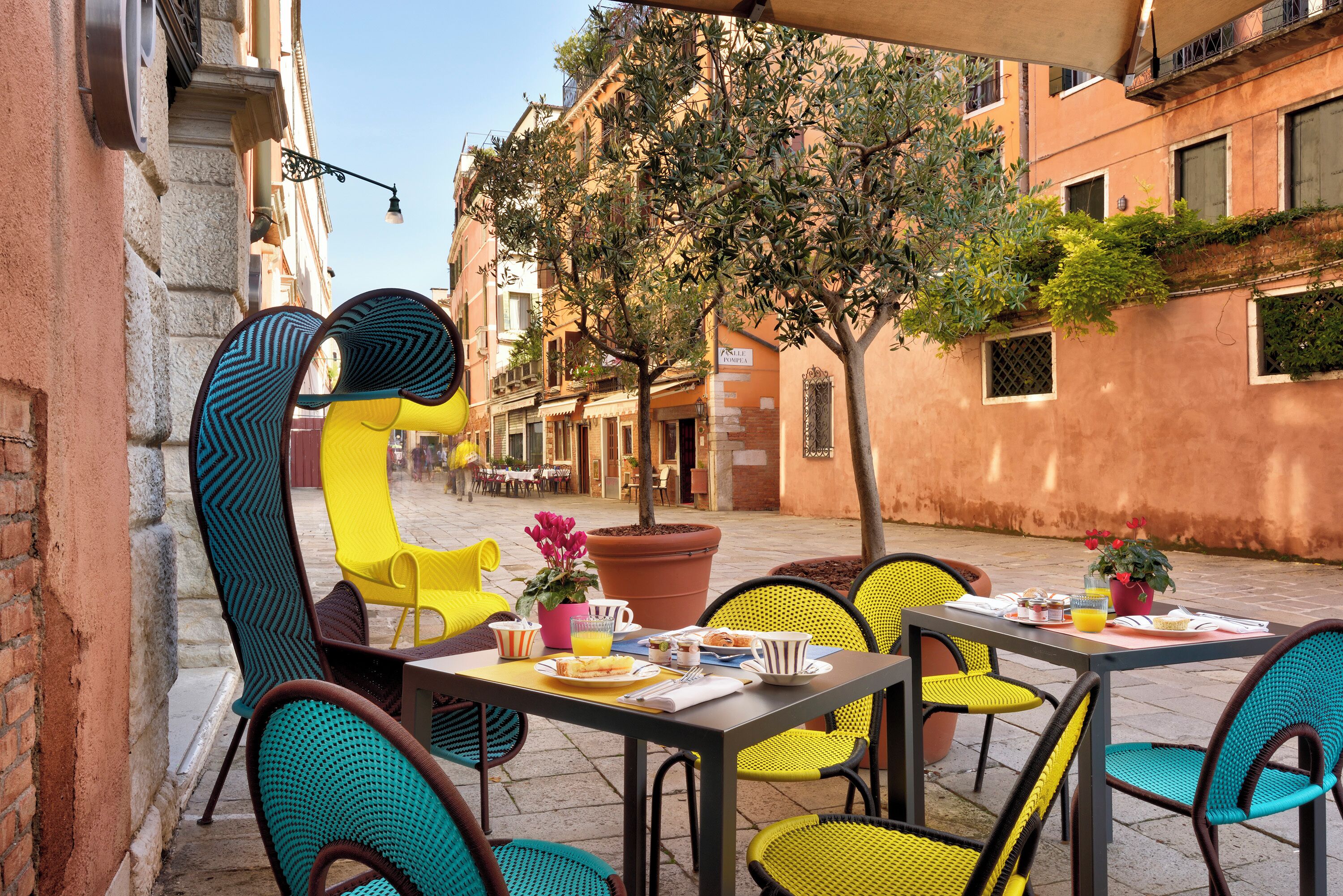An outdoor café with yellow and teal chairs, a table set with breakfast, and a potted plant.