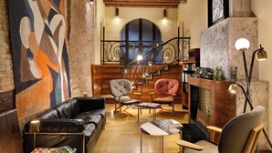 A cozy living room with a leather sofa, two armchairs, a coffee table, and a fireplace. A staircase with a wrought iron railing leads to an upper level.