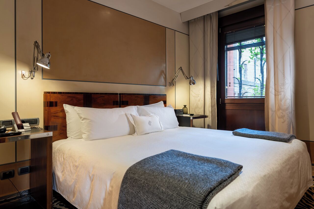 Une chambre d’hôtel avec un grand lit, deux tables de chevet, un bureau avec un téléphone et une fenêtre avec des stores.