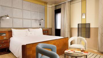 A hotel room with a large bed, a wooden nightstand, a blue chair, a small table with a tray of snacks, and a window with curtains.