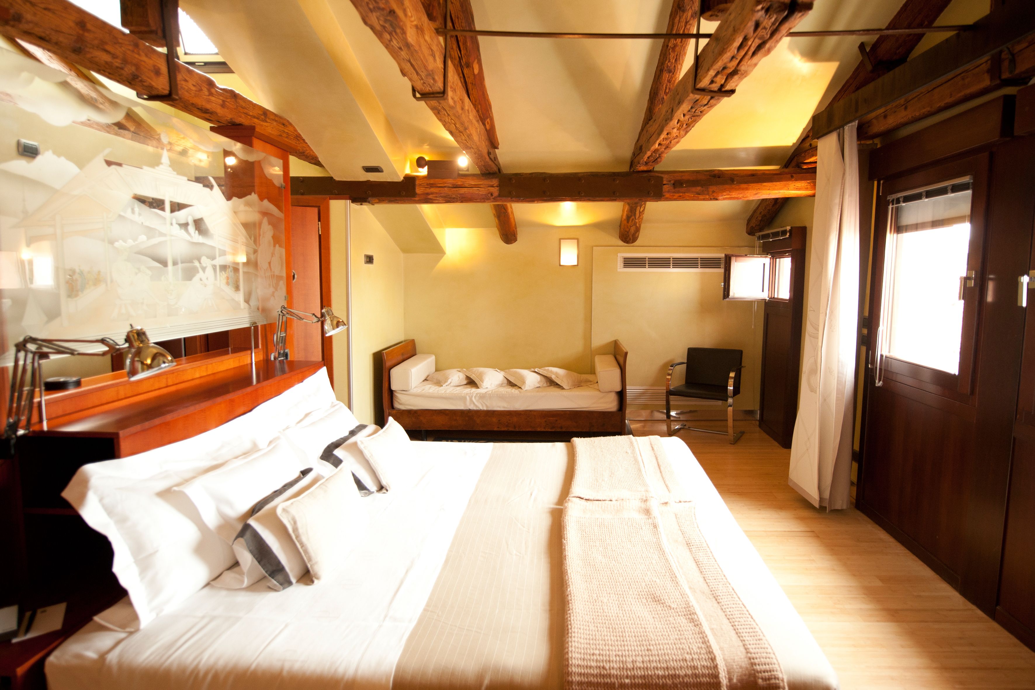 A hotel room with a large bed, a desk with a lamp, a chair, and a wooden ceiling.