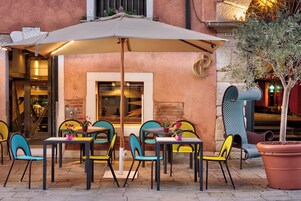 Zona all'aperto con tavoli, sedie, un ombrellone grande e una pianta in vaso.