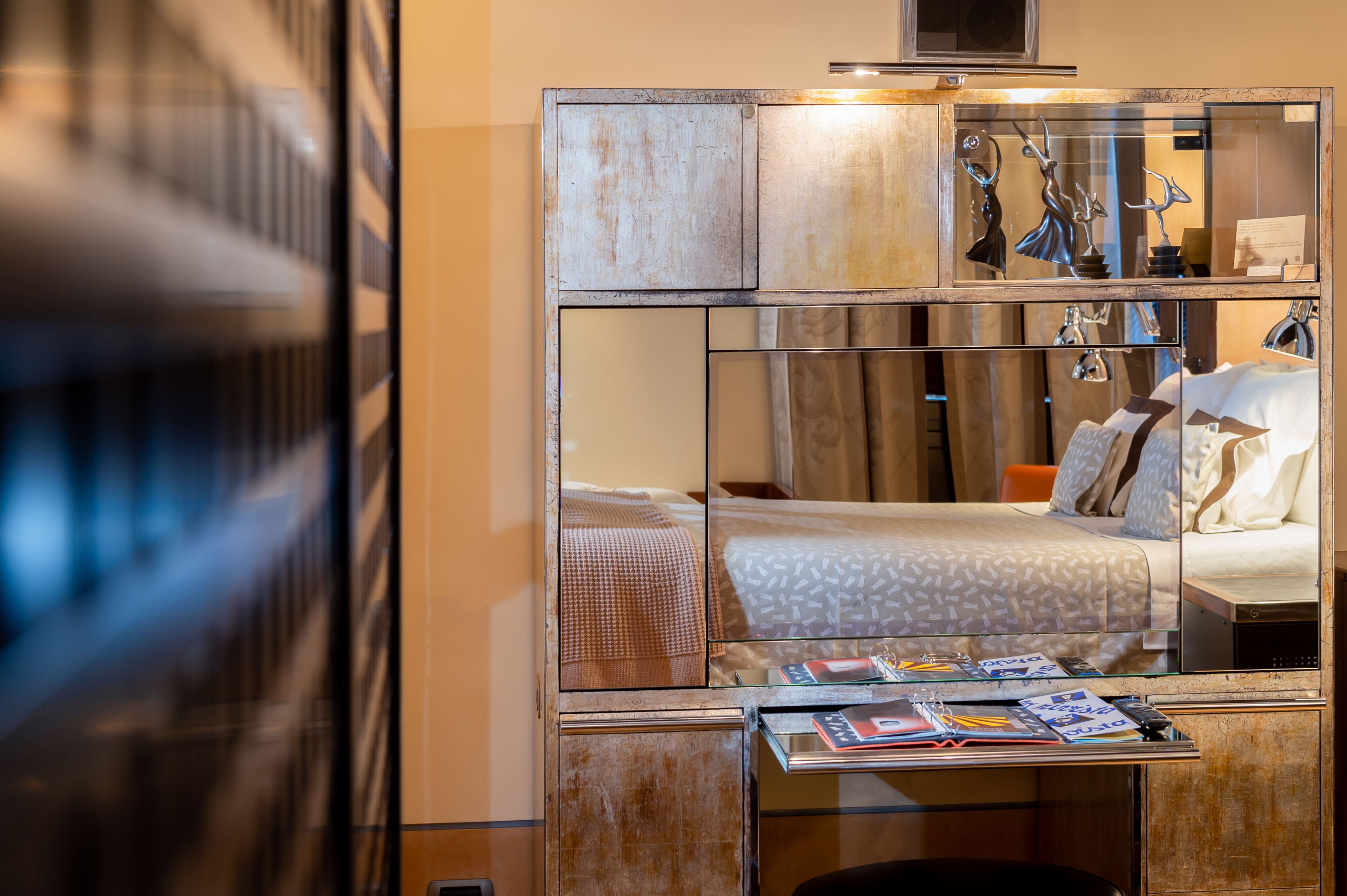 Ein kompaktes Hotelzimmer mit einem Spiegelkleiderschrank, einem Bett mit gemusterten Bettwaren und einem kleinen Tisch mit Zeitschriften.