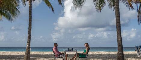 Zwei Personen sitzen an einem Tisch an einem sandigen Strand, im Hintergrund Palmen und das Meer.