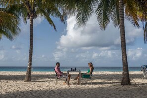 Dos personas sentadas en una mesa sobre una playa arenosa, con palmeras y el océano al fondo.