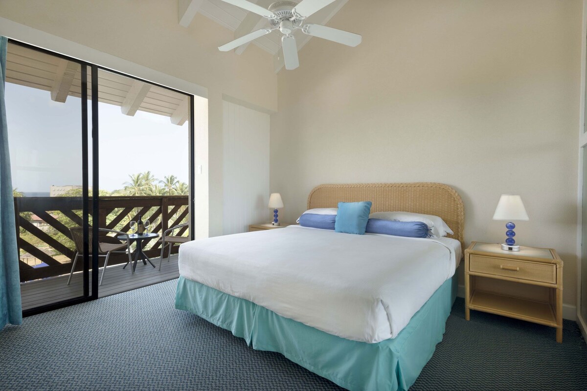 Habitación de hotel con cama, dos mesitas de noche, ventilador de techo y balcón con vistas a palmeras.