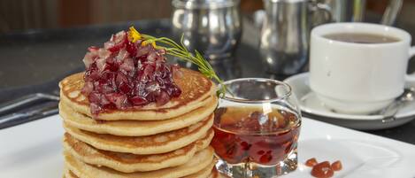 Una pila de tortitas con dulce de frutos rojos y jarabe, un vaso pequeño de jarabe y una taza de café sobre un plato blanco.
