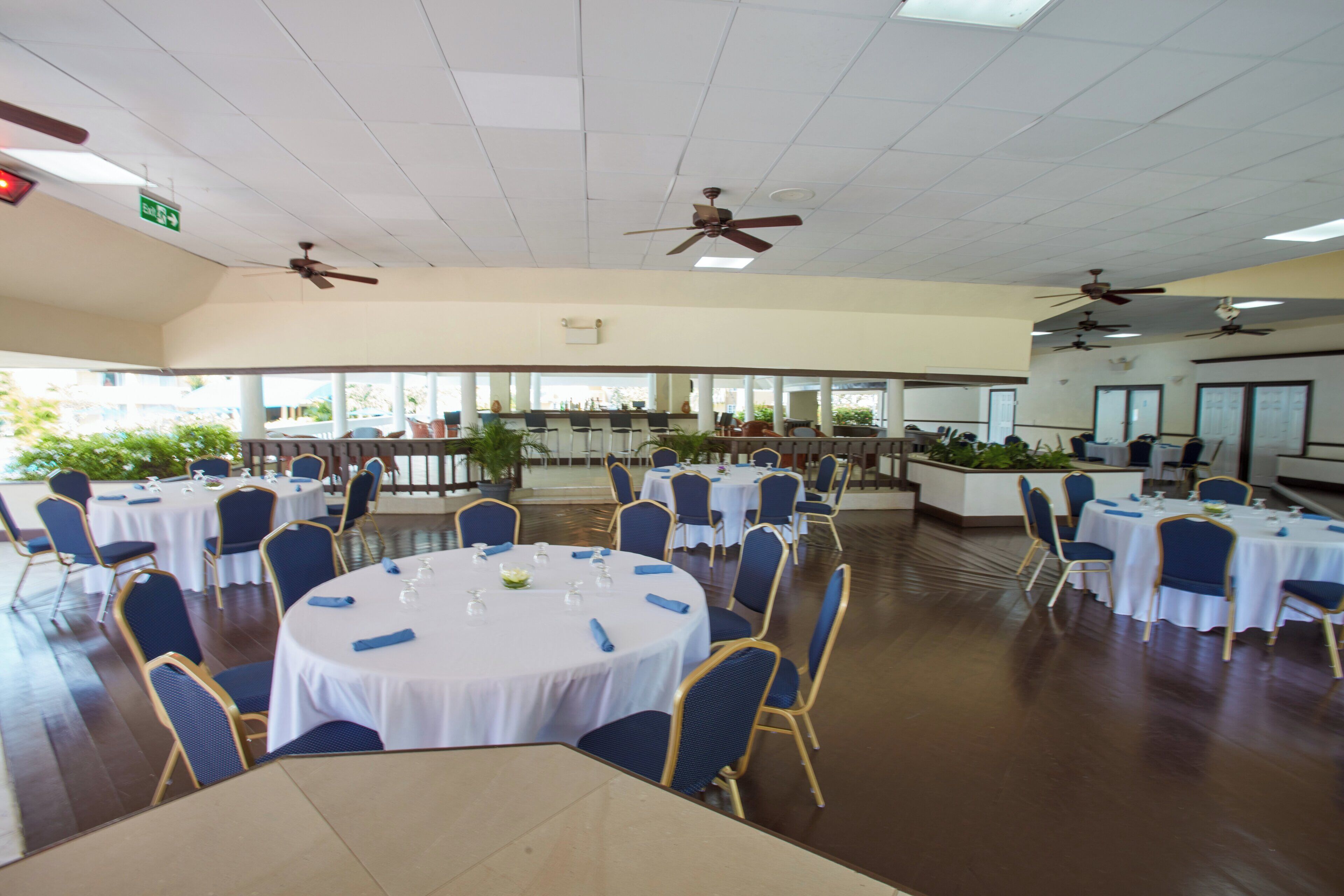 A spacious indoor dining area with round tables, blue chairs, ceiling fans, and large windows.