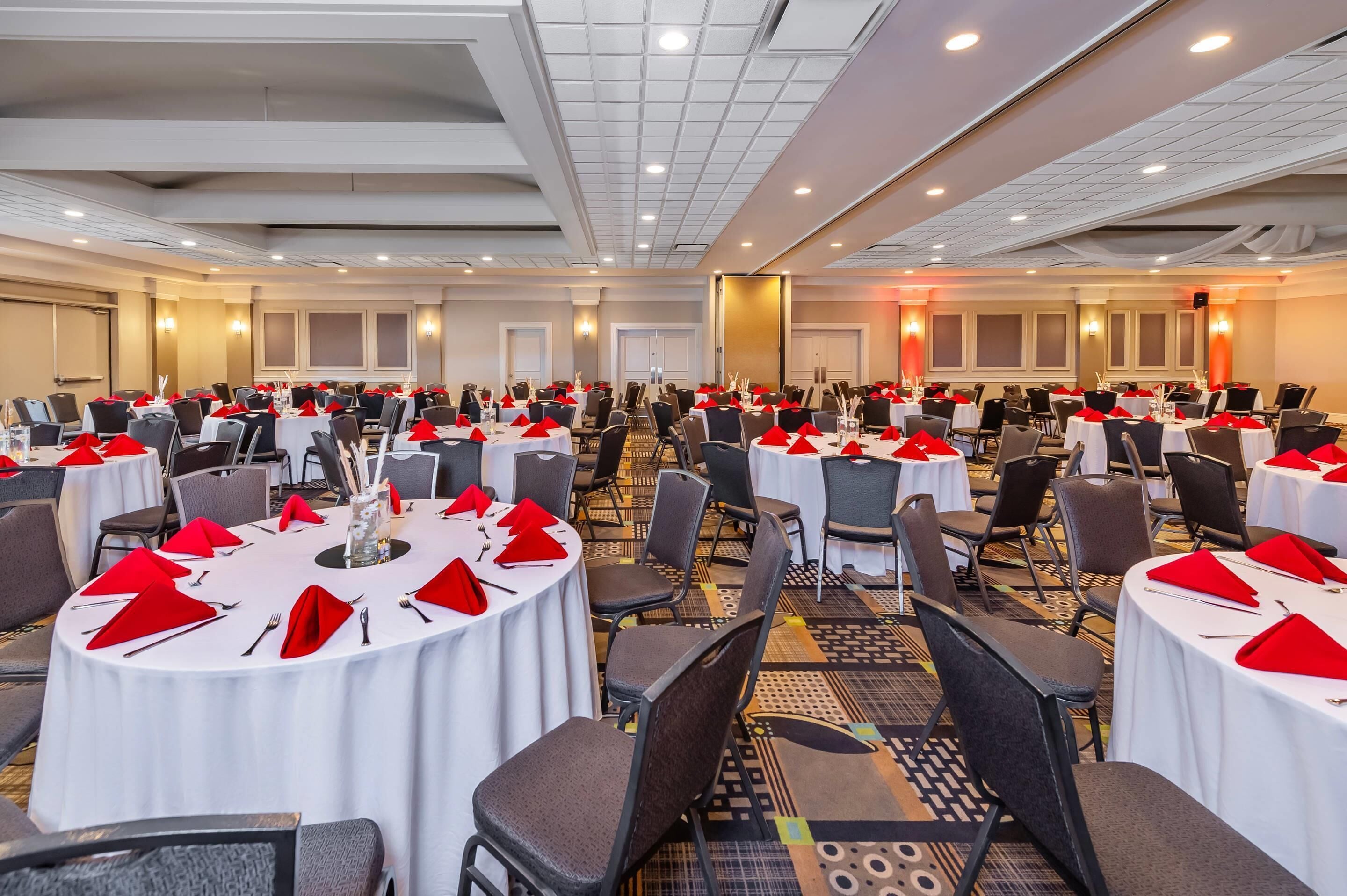 Une grande salle de conférence équipée de plusieurs tables rondes, chacune agrémentée de serviettes rouges et de couverts en argent.