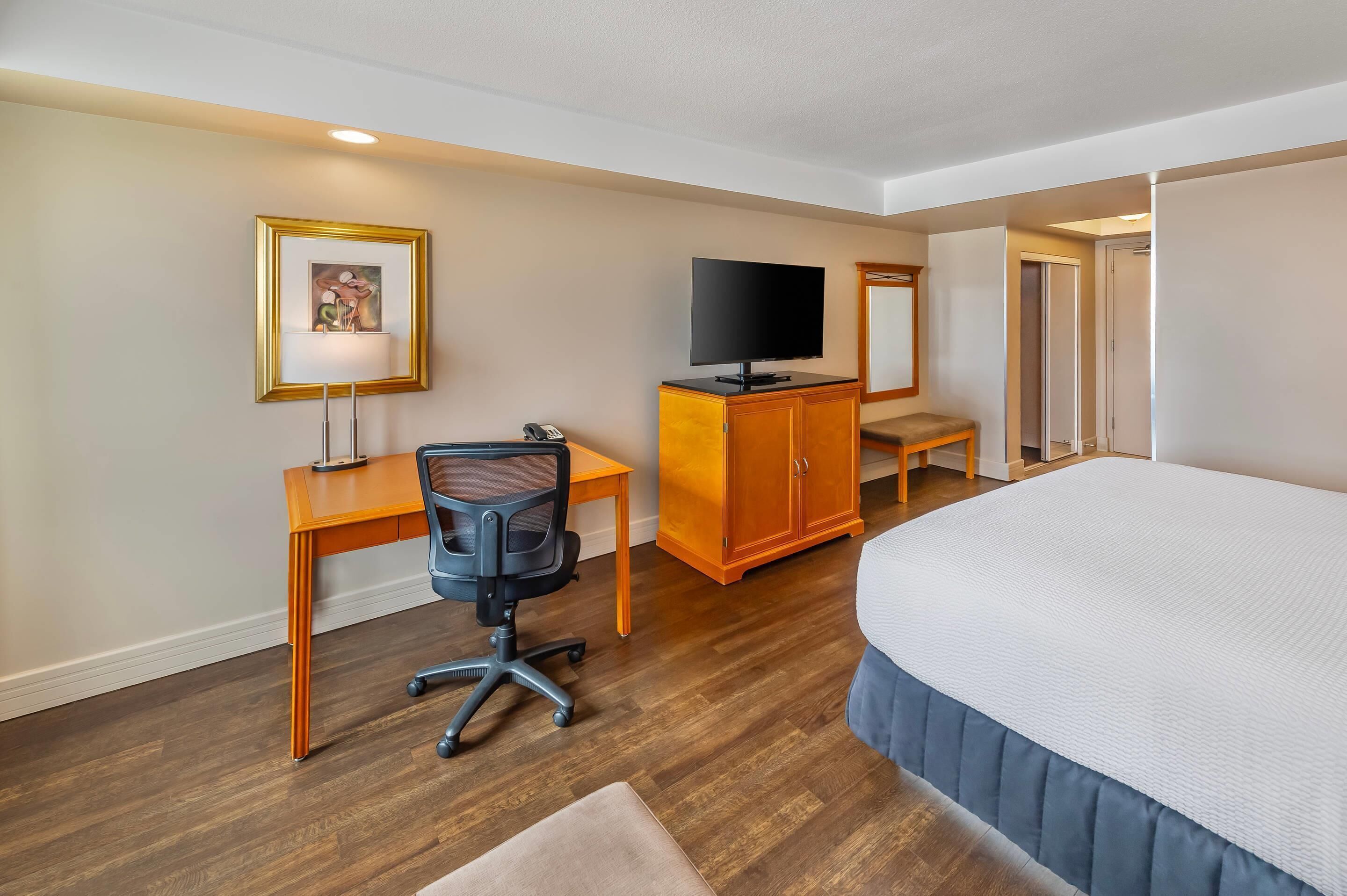 A hotel room with a bed, a desk with a chair, a television, a cabinet, and a framed picture on the wall.