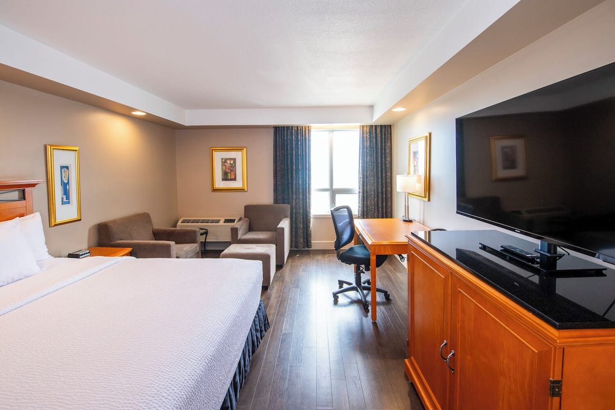 A hotel room with a bed, a desk with a television, a chair, a sofa, and a window with curtains.