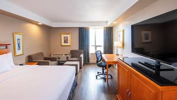 A hotel room with a bed, a desk with a television, a chair, a sofa, and a window with curtains.