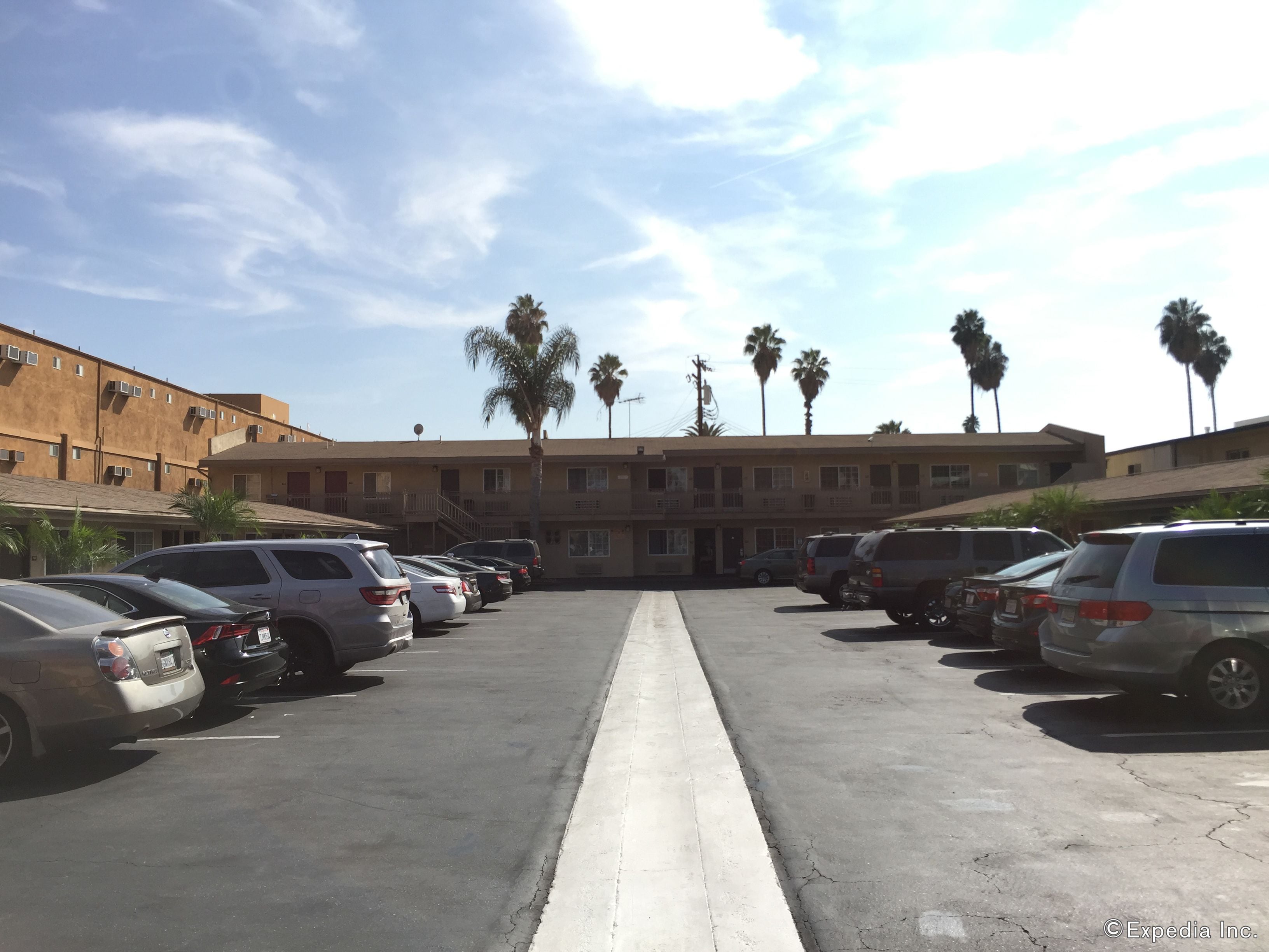 A parking lot with multiple cars, a building with balconies, and palm trees in the background.