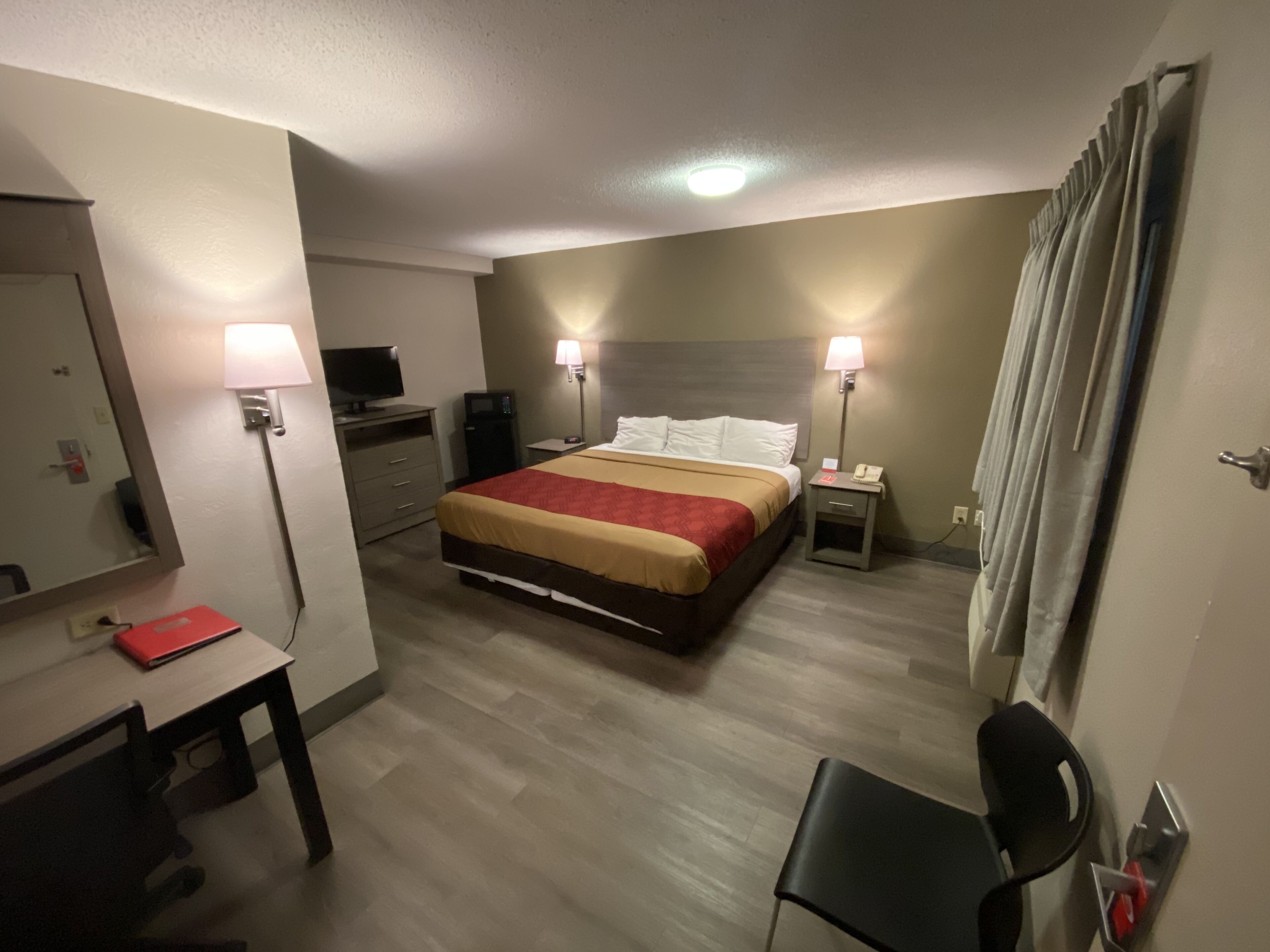 A hotel room with a bed, bedside lamps, a desk with a red folder, a TV, a mirror, and a chair.