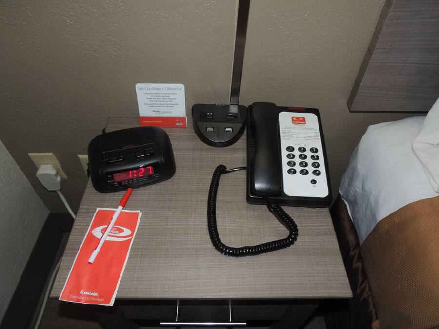 Habitación de hotel con un escritorio que contiene un teléfono negro, un reloj digital y una pluma roja.
