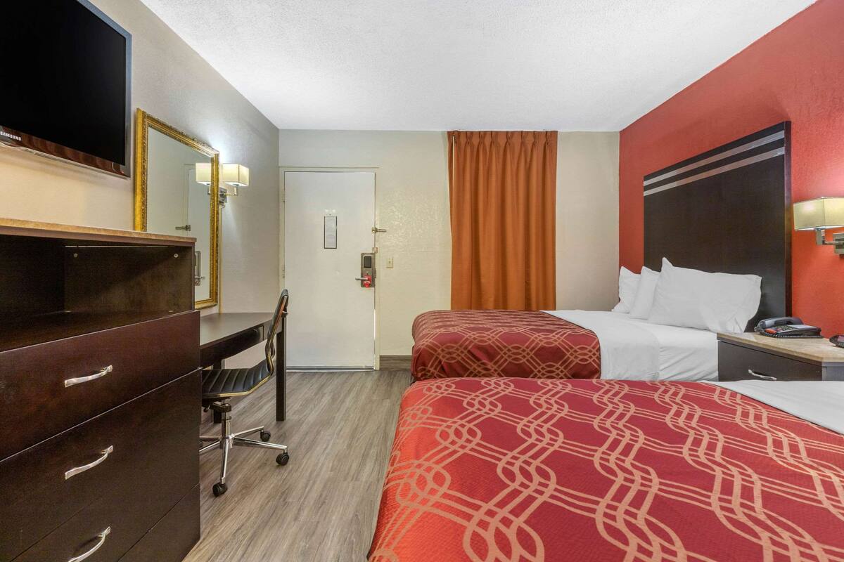 A hotel room with two beds, a desk with a chair, a television, and a door with a security panel.