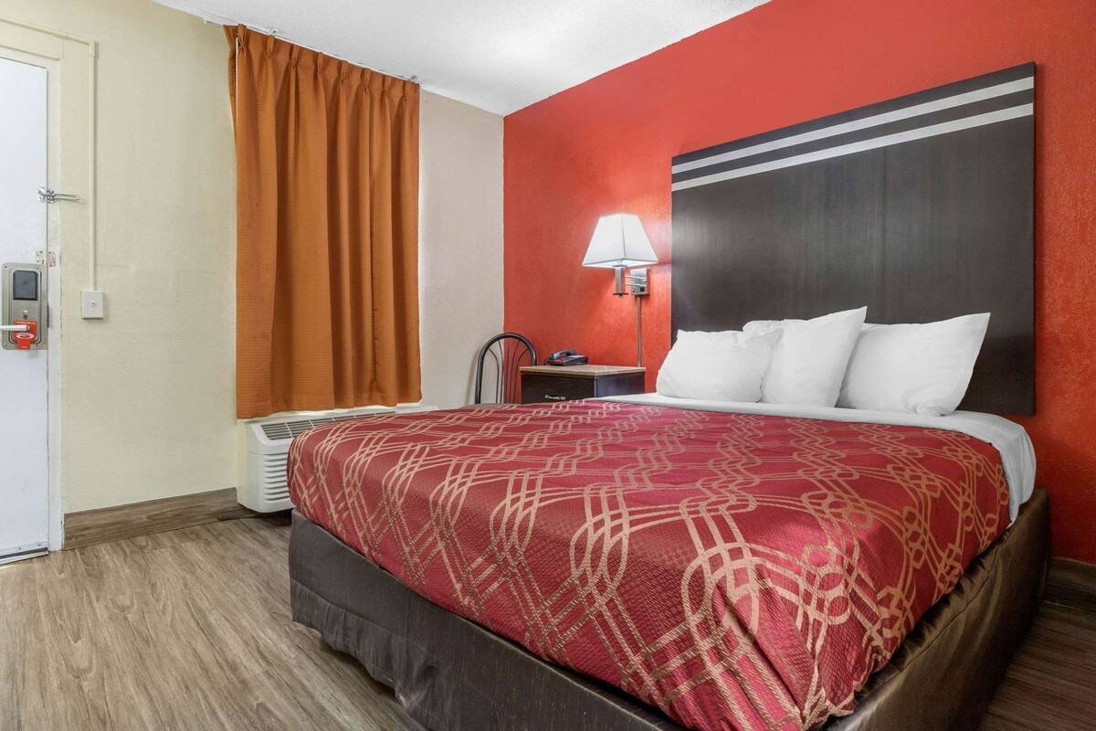 A hotel room with a bed, a nightstand, a lamp, a heater, and a door.