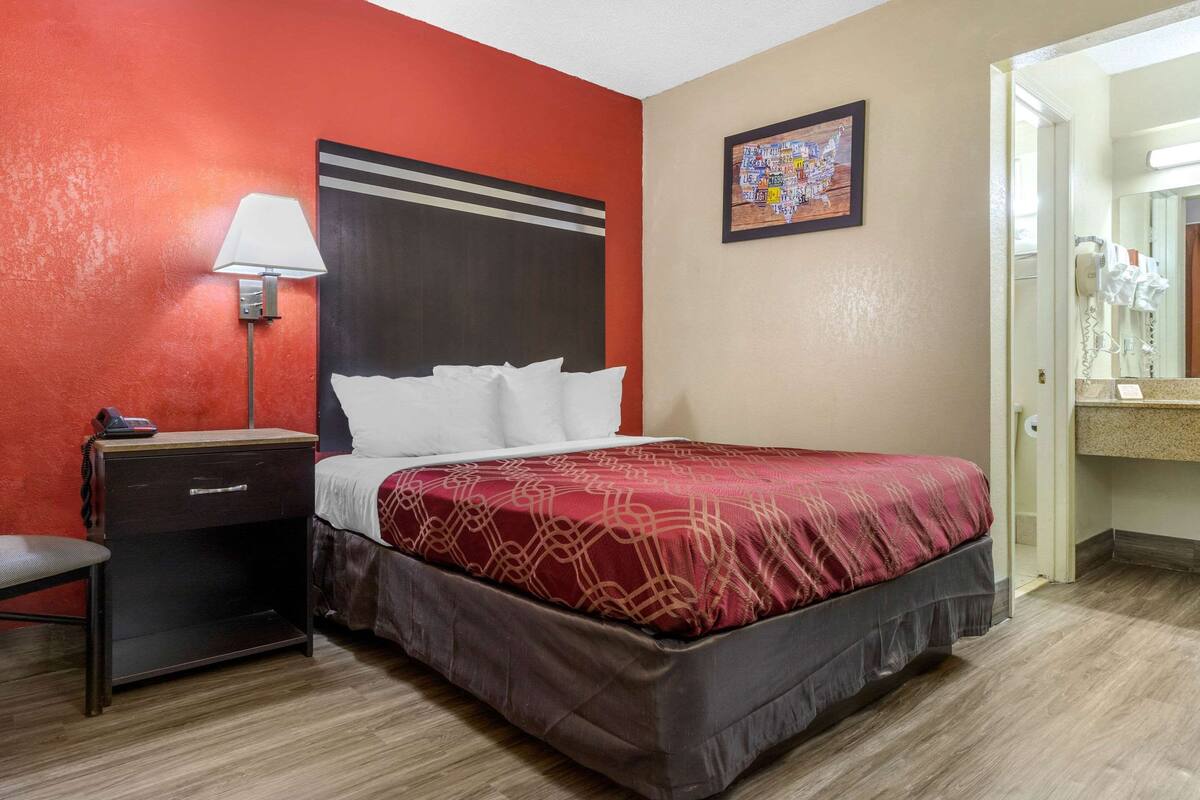 A hotel room with a bed, a nightstand, a lamp, a painting, and a bathroom visible through an open door.