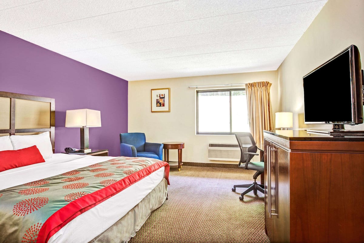 A hotel room with a large bed, a desk with a television, a chair, a small table, and a window with curtains.