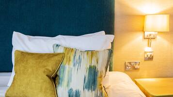 Una cama bien tendida con dos almohadas, una lisa y otra estampada, en una habitación con cabecera azul y una lámpara de pared.