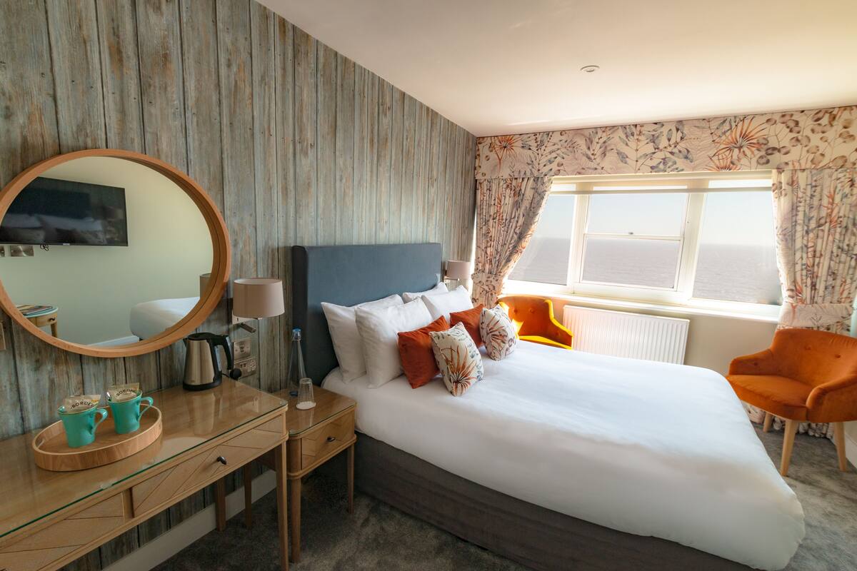 Habitación de hotel con una cama grande, un aparador de madera, un espejo, una ventana con cortinas y una silla.