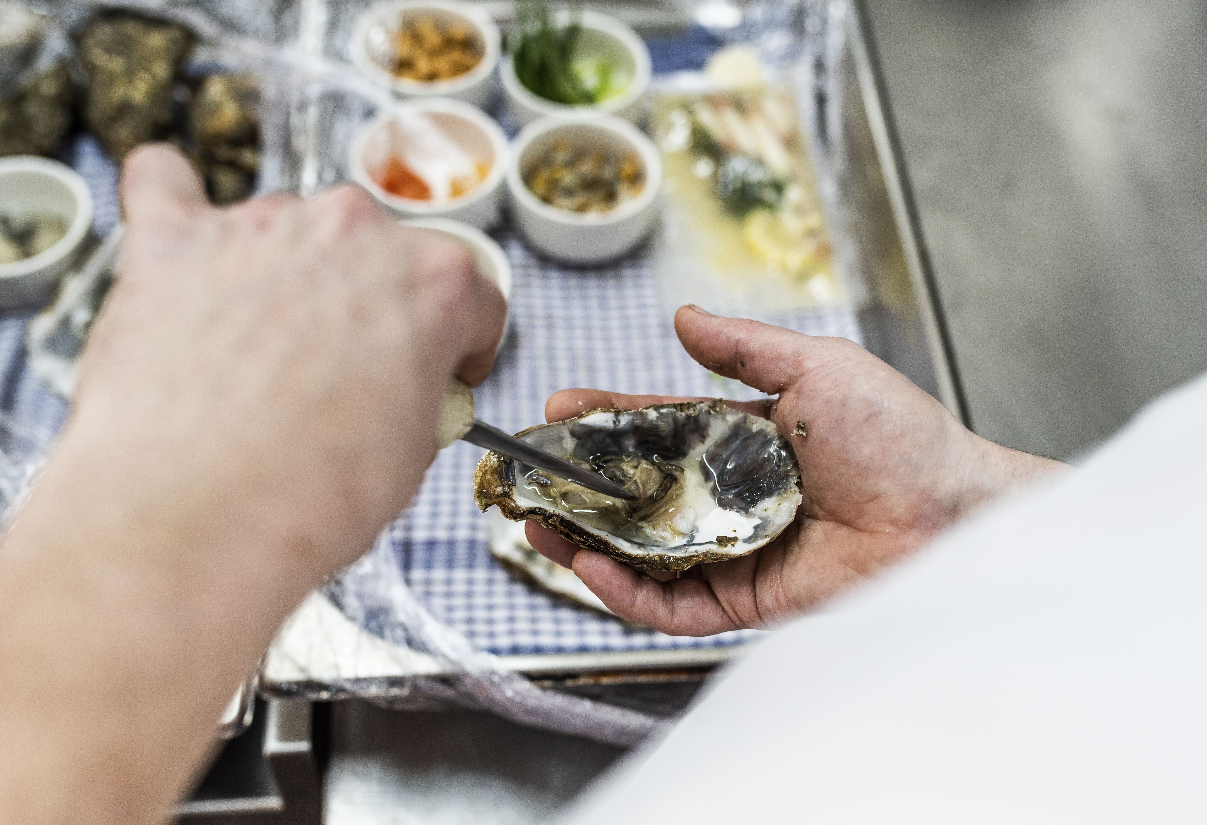 A person holding an oyster with a knife, surrounded by various condiments on a checkered tablecloth.