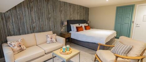 Quarto com cama, cabeceira de madeira, mesa de cabeceira, sofá bege, poltrona de madeira e uma pequena mesa com bandeja.