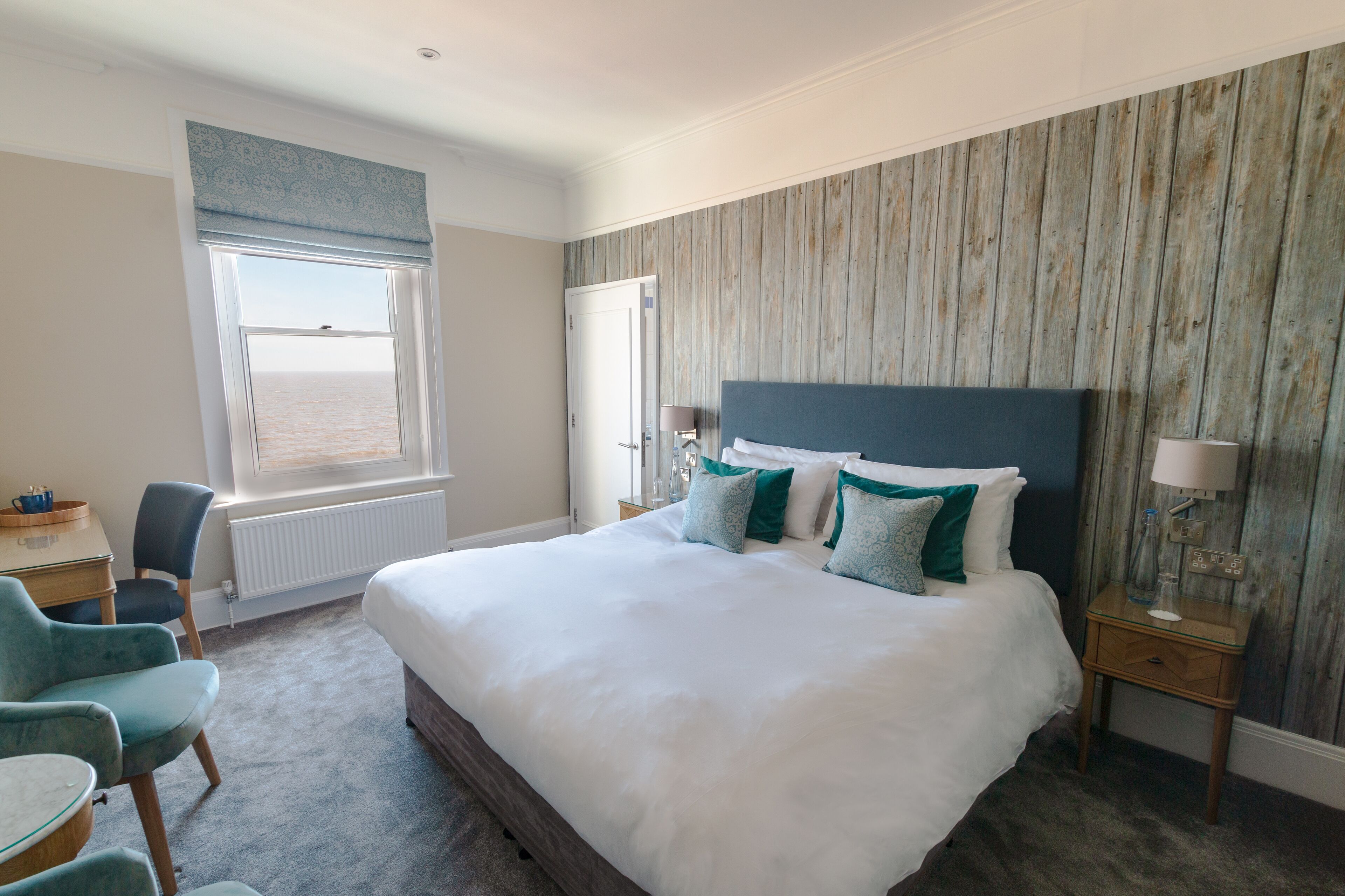 Un dormitorio con una cama grande, un escritorio y una silla. Se ve una ventana con vistas al mar.