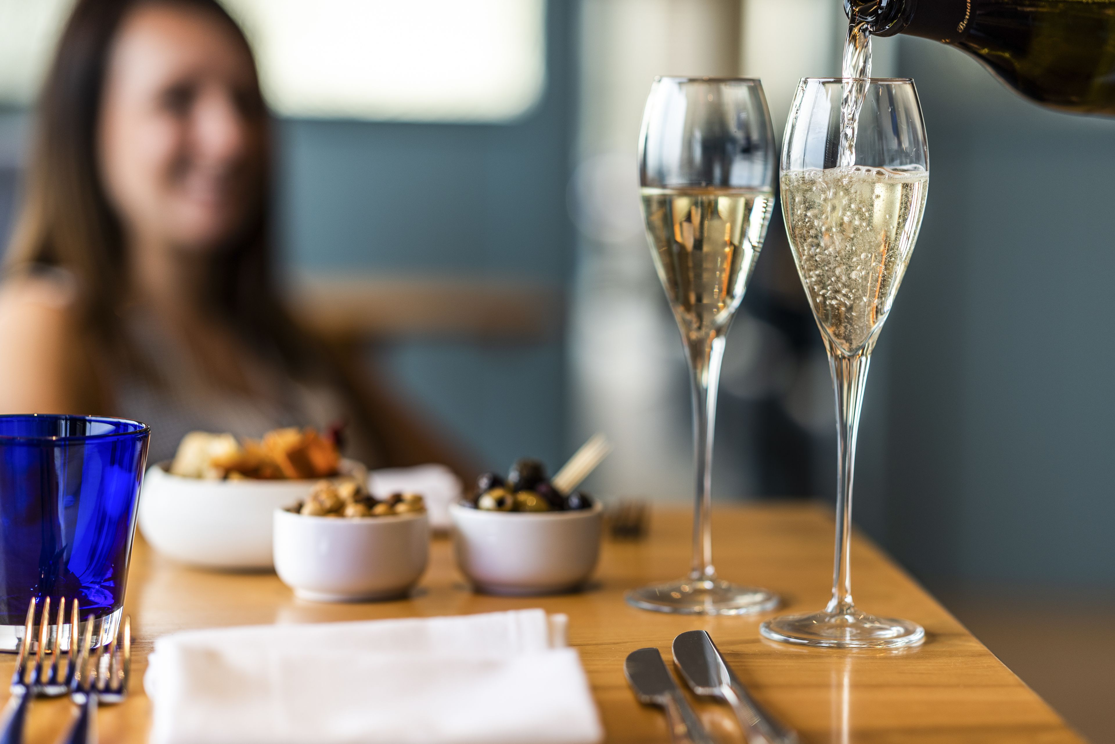A dining table with a bottle of champagne being poured into two glasses, bowls of olives and nuts, and a blue glass.
