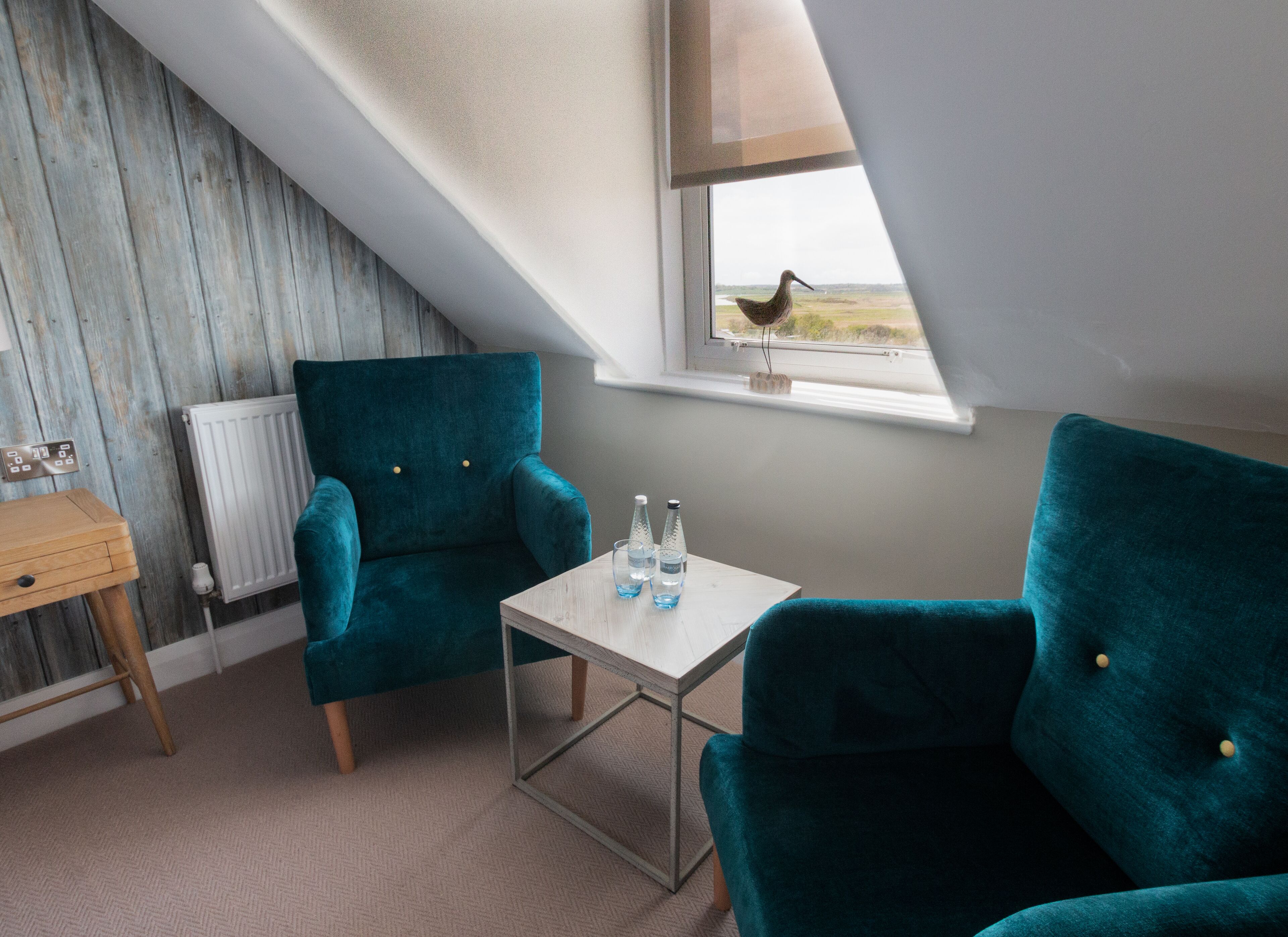 A room with teal chairs, a small table, a radiator, and a window with a view of the beach.