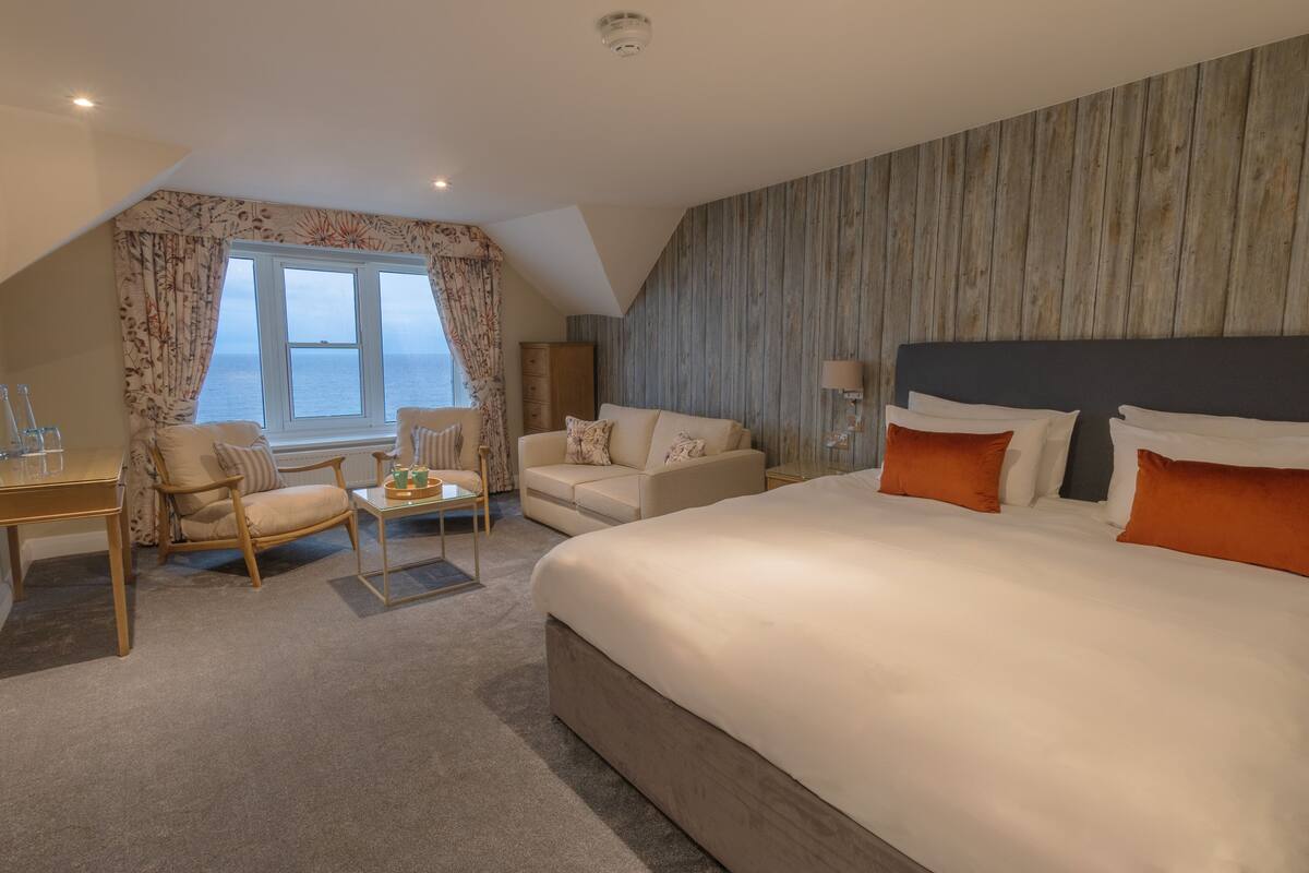 Habitación de hotel con una cama grande, un sofá, una mesita pequeña y una silla. Tiene una ventana con cortinas, una pared de madera y una lámpara en la mesita de noche.