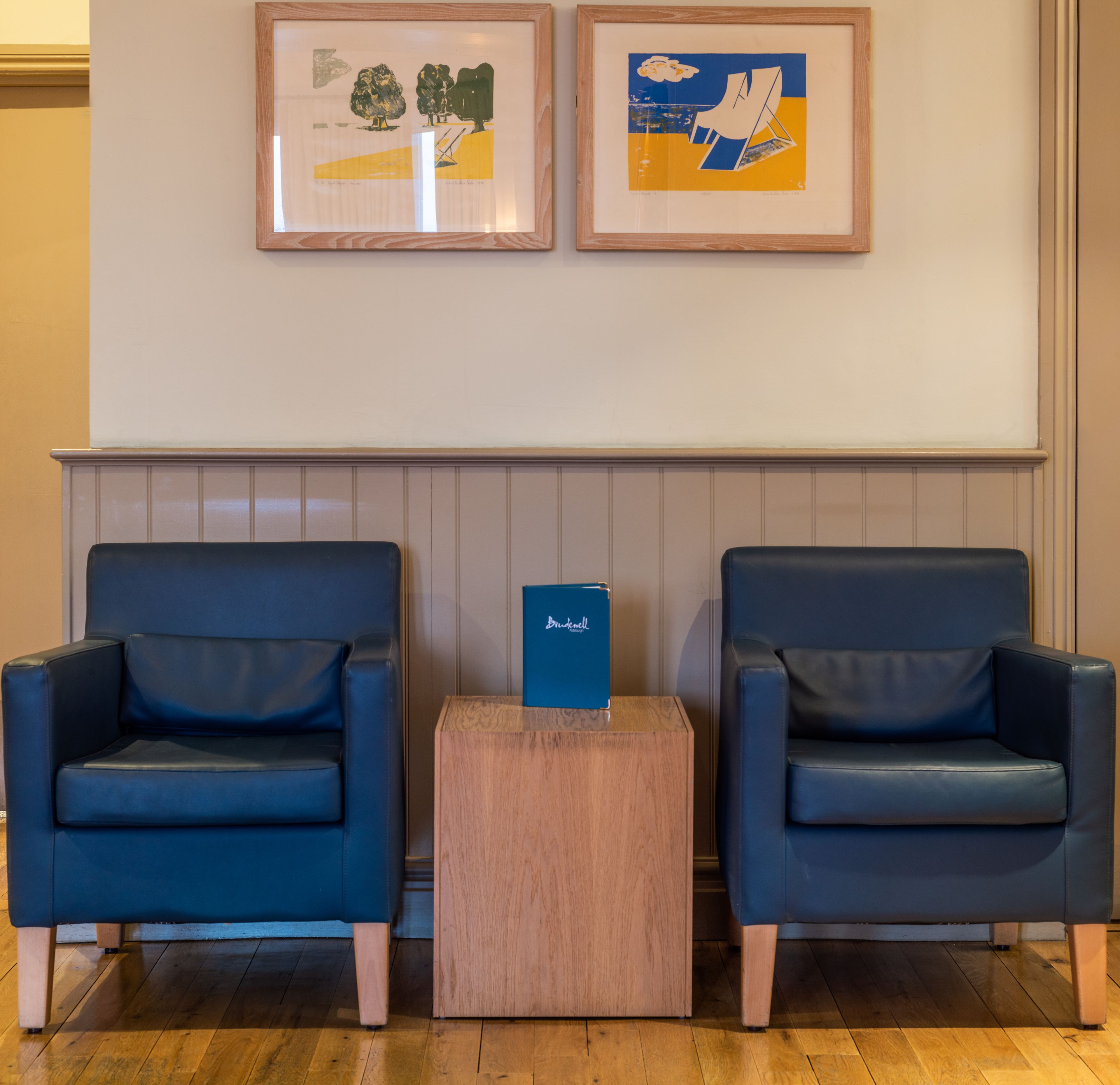 Deux fauteuils bleus, séparés par une petite table d’appoint en bois, une œuvre d’art encadrée au-dessus, et un mur lambrissé.
