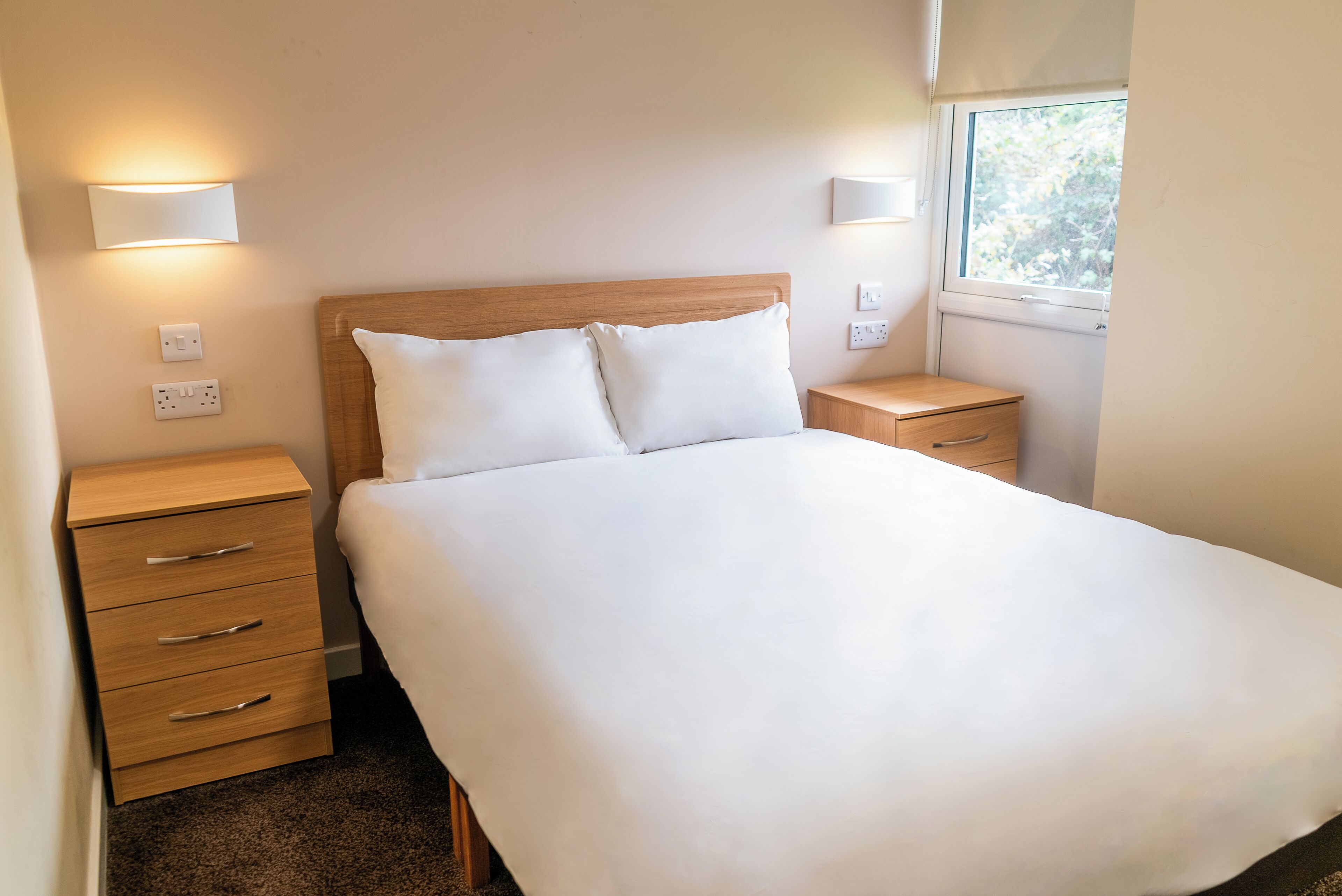Cama bem arrumada com roupa de cama branca, cabeceira de madeira e duas mesas de cabeceira com abajures fixados na parede.