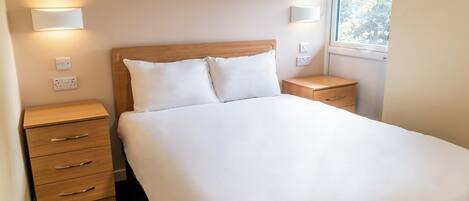 Cama bem arrumada com roupa de cama branca, cabeceira de madeira e duas mesas de cabeceira com abajures fixados na parede.