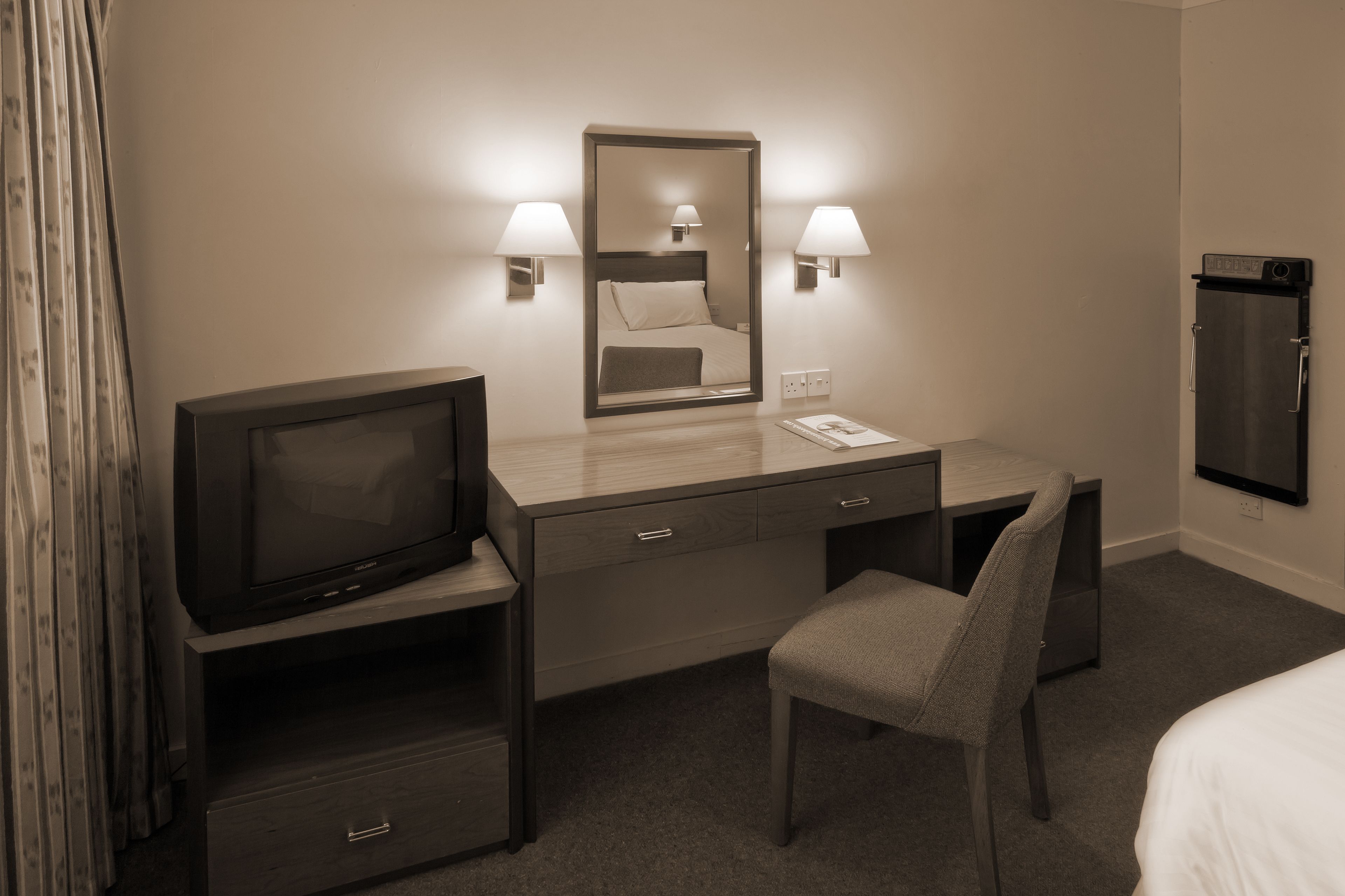 A hotel room with a bed, a desk with a television, a chair, a mirror, and a refrigerator.