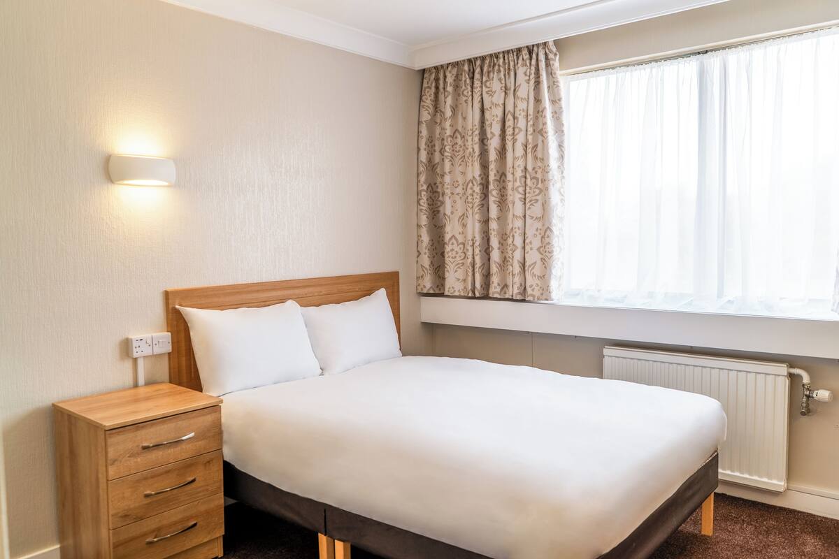 Een hotelkamer met een houten bed, een nachtkastje met lades, een wandlamp, een raam met gordijnen en een radiator.