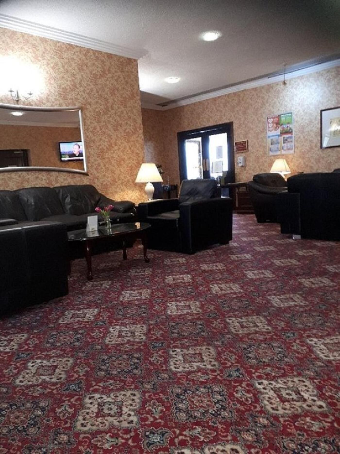 Un vestíbulo de hotel con una alfombra roja estampada, sofás de cuero negro, una mesa de centro y un televisor en la pared.