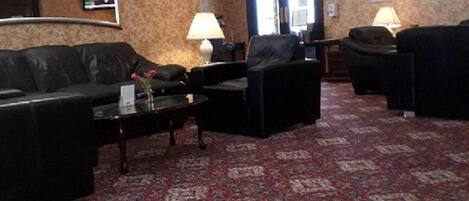 Un vestíbulo de hotel con una alfombra roja estampada, sofás de cuero negro, una mesa de centro y un televisor en la pared.