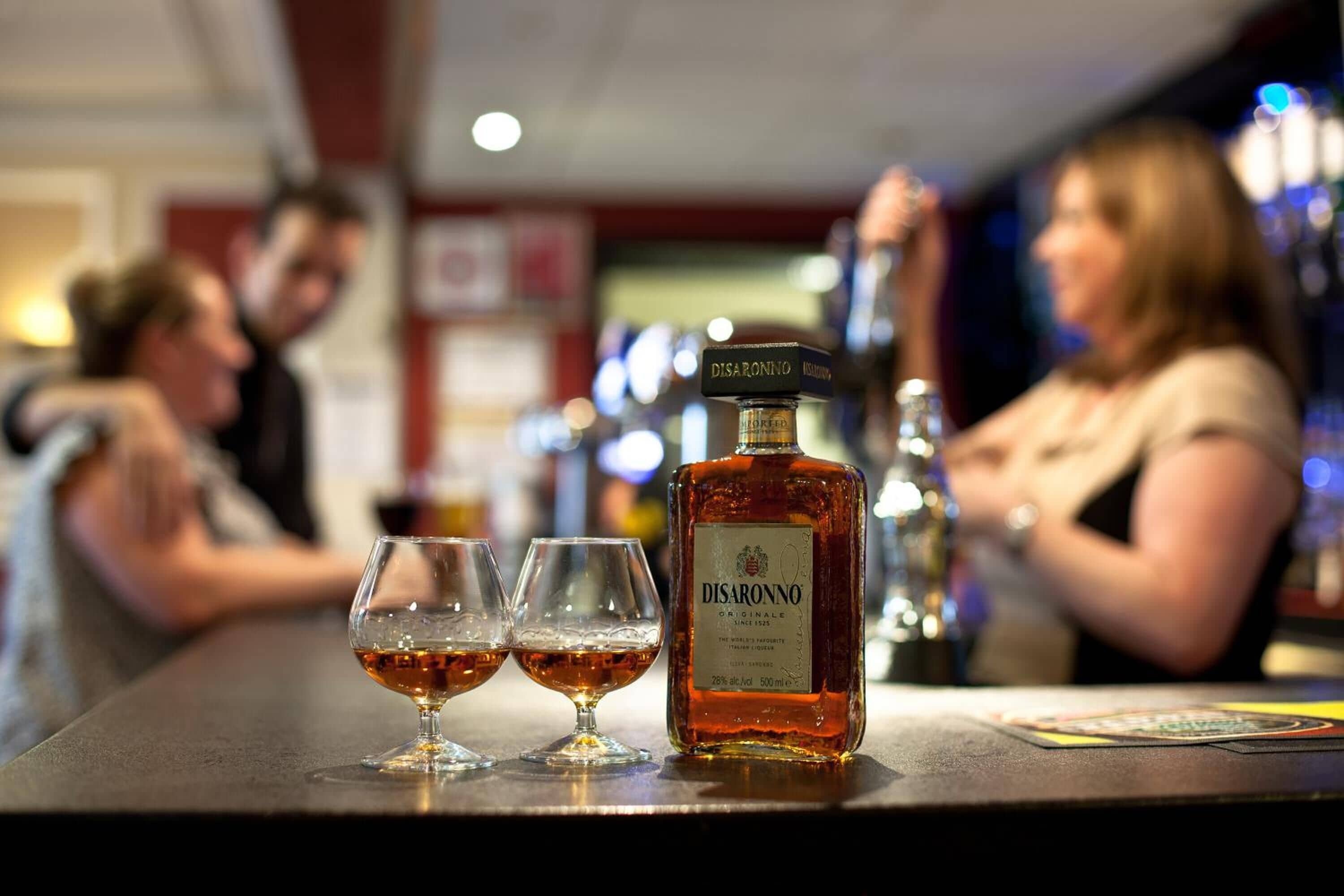 A bar setting with a bottle of Disaronno on the counter, two glasses of the beverage, and a blurred background of people.