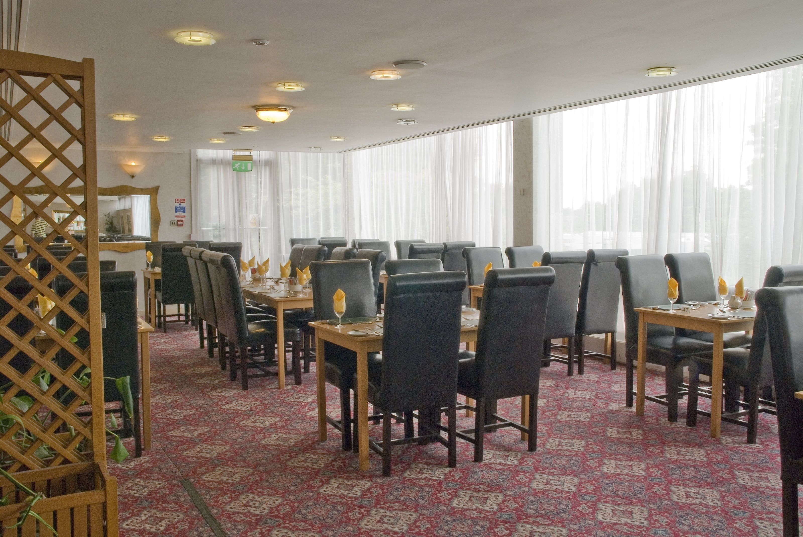 A dining area with tables and chairs, large windows with sheer curtains, and a patterned carpet.