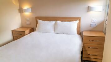 Una cama bien hecha con sábanas blancas, dos mesitas de noche con cajones e lámparas fijadas a la pared.