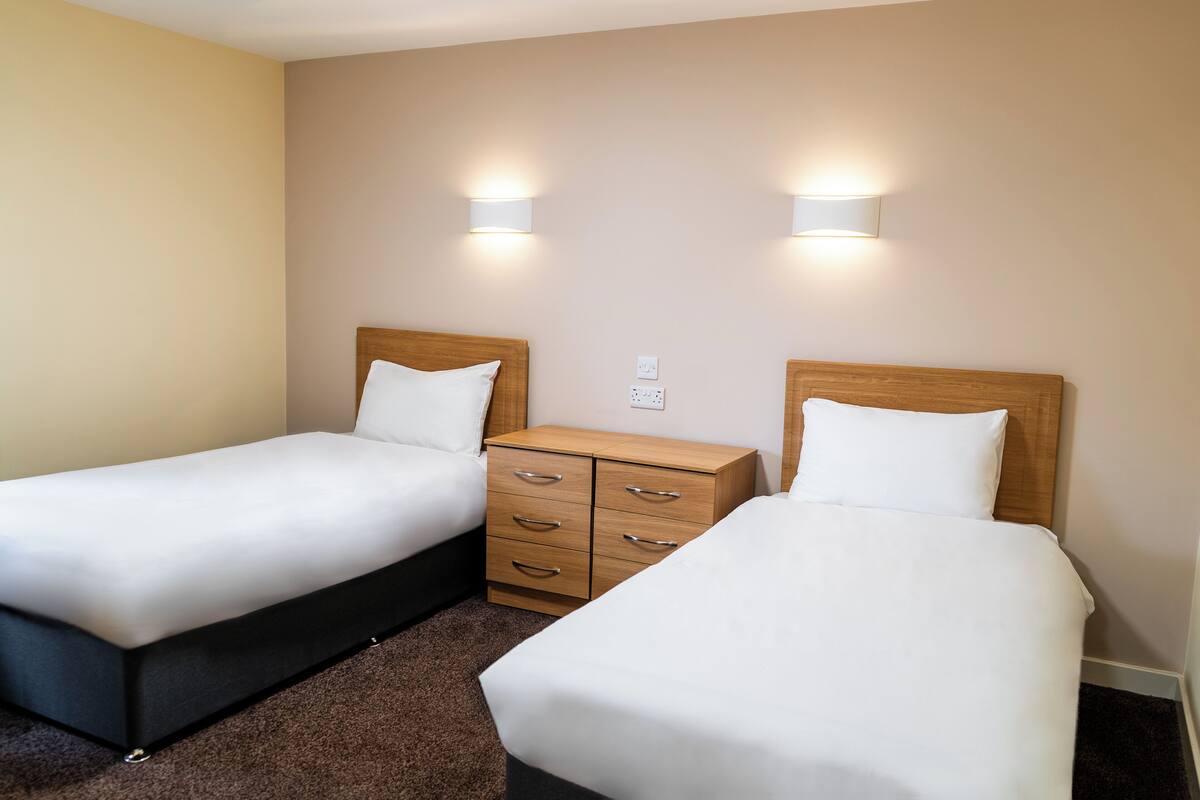 Twee enkele bedden met houten hoofdborden, een klein houten dressoir en twee wandlampen in een hotelkamer.