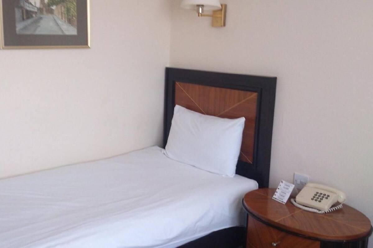 Een eenpersoonsbed met een houten hoofdbord, een nachtkastje met een telefoon, en een ingelijste foto aan de muur.