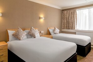 Een hotelkamer met twee bedden, elk met een houten hoofdbord, witte beddengoed en decoratieve kussens. Er is een nachtkastje met een lamp en een stopcontact op de muur.