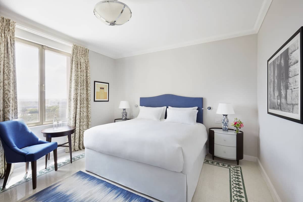 A hotel room with a large bed, a blue armchair, a small table, a nightstand, and a view of the city.