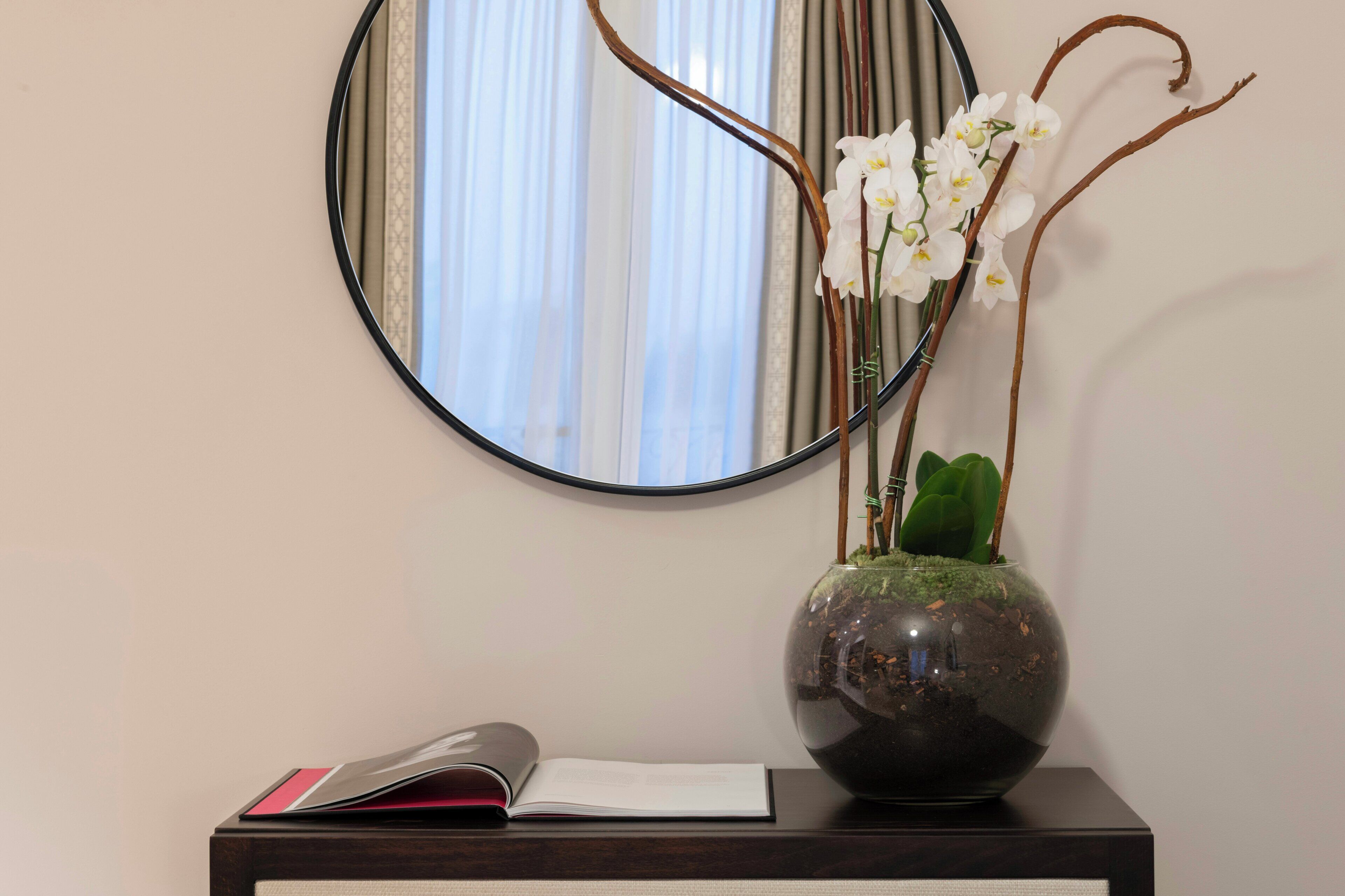 A desk with a vase of white orchids, a book, and a mirror reflecting a window with curtains.